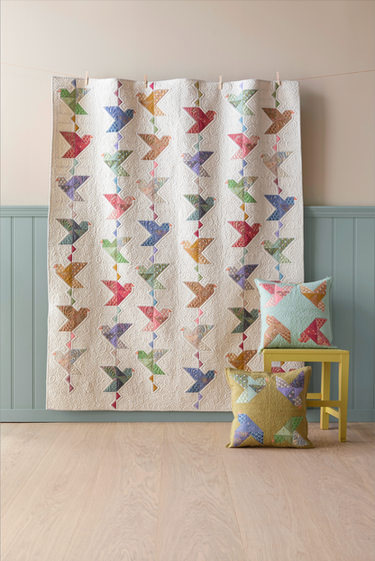 A quilted fabric panel or curtain featuring a repeating pattern of colorful birds on an off-white background, hanging in a room with a light wood floor, pale wall, and blue-green wainscoting. Two matching decorative pillows rest on a small yellow stool.
Photo by Tildafabrics.com [additional_image_link]