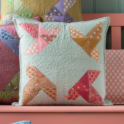A completed square-shaped pillow with a paperbird motif, featuring a combination of sage green and other colors, placed on a pink bench with various other fabric pillows.
Photo by Tildafabrics.com [image_link]
