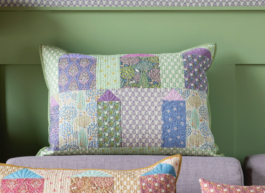 A patchwork pillow featuring a repeating pattern of small house-shaped blocks in pastel green, lavender, purple, blue, and cream.
Photo by Tildafabrics.com [image_link]