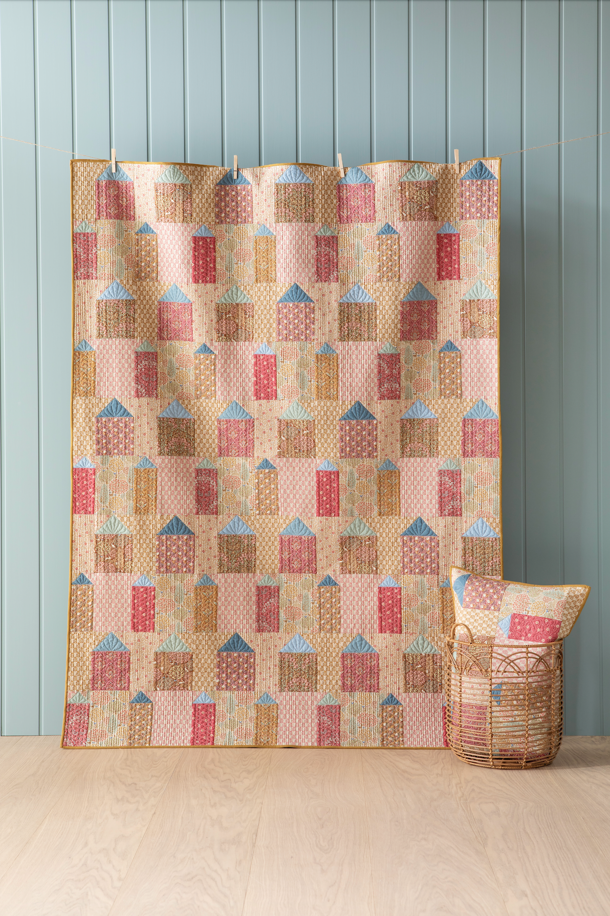 A quilt with a geometric pattern featuring small houses in warm colors, hanging on a wall with a blue wall panel and a small decorative pillow to the side.
Photo by Tildafabrics.com [image_link]