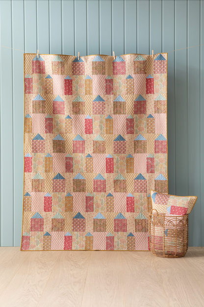 A quilt with a geometric pattern featuring small houses in warm colors, hanging on a wall with a blue wall panel and a small decorative pillow to the side.
Photo by Tildafabrics.com [image_link]