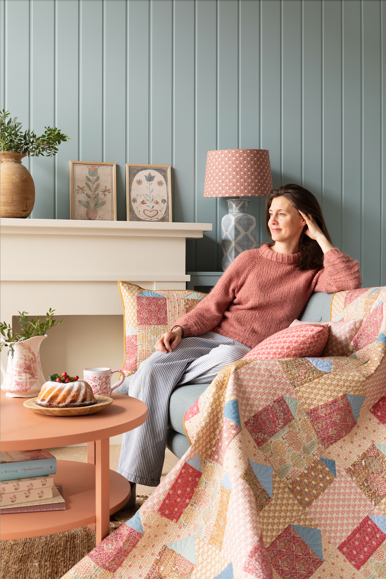 A quilt with a geometric pattern featuring small houses in warm colors, hanging on a wall with a blue wall panel and a small decorative pillow to the side.
Photo by Tildafabrics.com [additional_image_link]