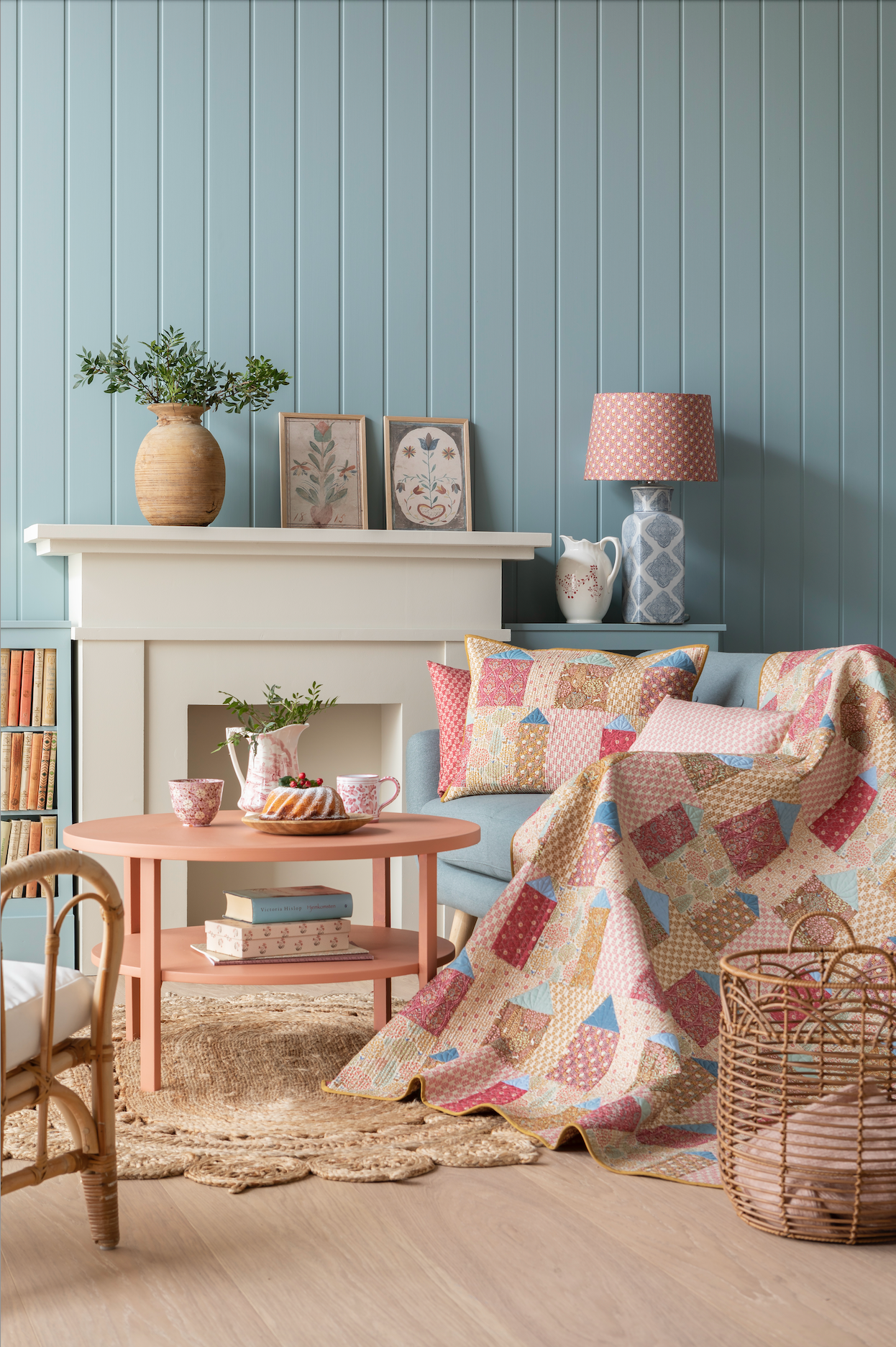 A quilt with a geometric pattern featuring small houses in warm colors, hanging on a wall with a blue wall panel and a small decorative pillow to the side.
Photo by Tildafabrics.com [additional_image_link]