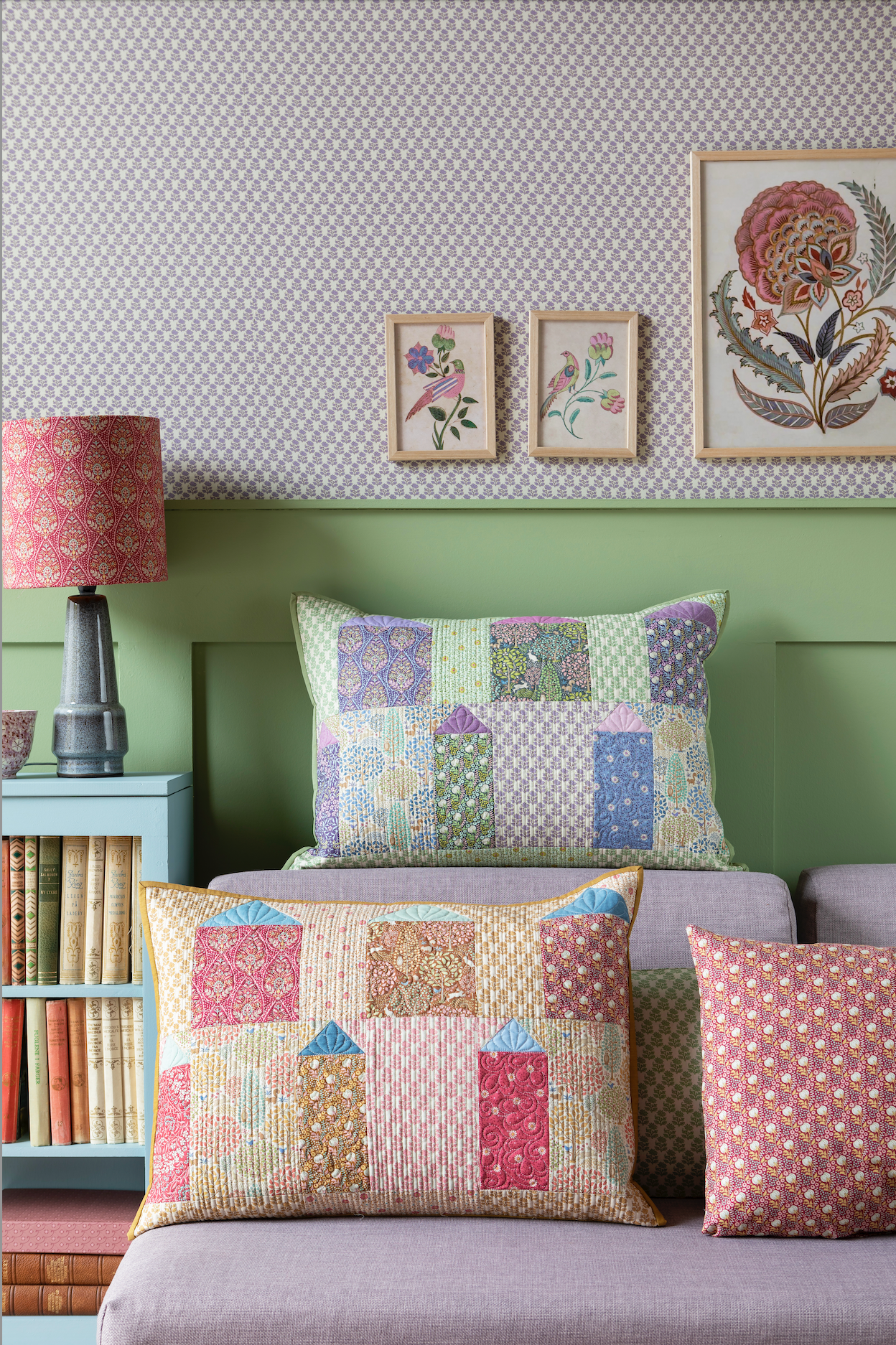 A quilt with a geometric pattern featuring small houses in warm colors, hanging on a wall with a blue wall panel and a small decorative pillow to the side.
Photo by Tildafabrics.com [additional_image_link]