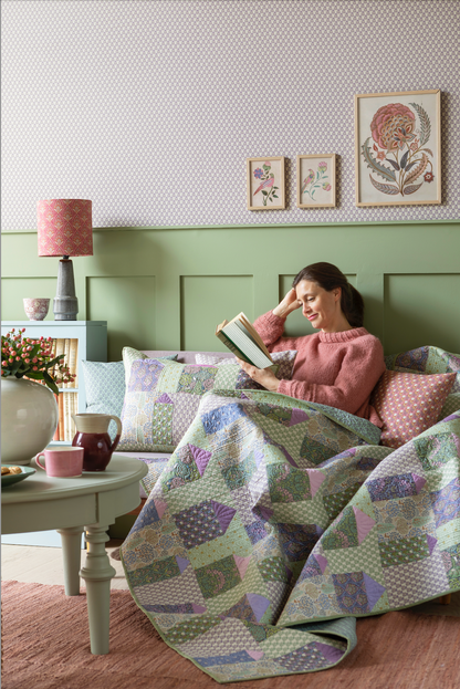 A quilt with a geometric pattern featuring small houses in cool colors, hanging on a wall with a blue wall panel and a small decorative pillow to the side.
Photo by Tildafabrics.com [additional_image_link]