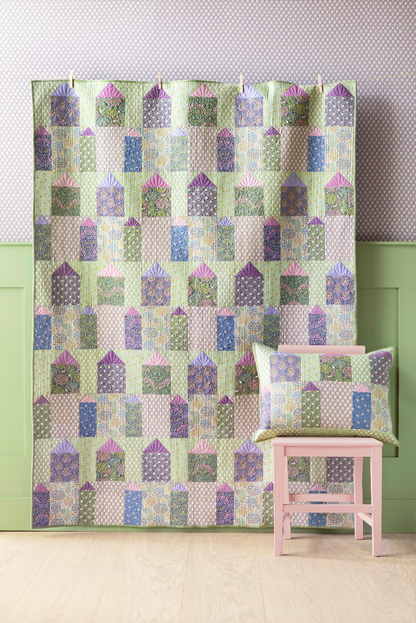 A quilt with a geometric pattern featuring small houses in cool colors, hanging on a wall with a blue wall panel and a small decorative pillow to the side.
Photo by Tildafabrics.com [additional_image_link]