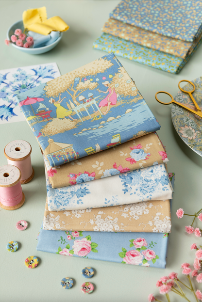 A stack of folded fabric pieces with pastel floral and scenic prints, arranged on a light surface with sewing props including spools of thread, scissors, buttons, and additional patterned fabric.
Photo by Tildafabrics.com [additional_image_link]