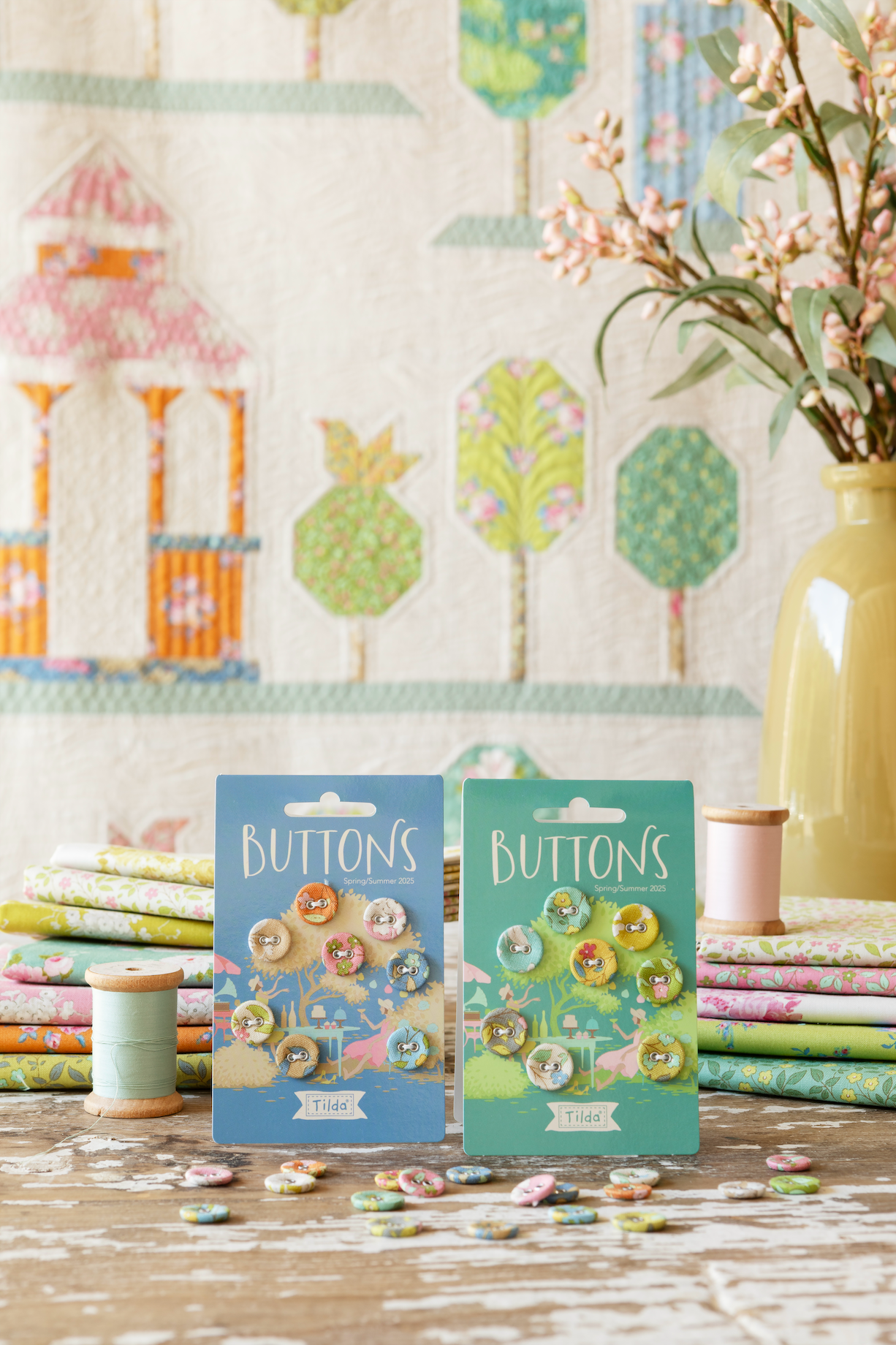 Two packages of decorative buttons displayed on a rustic wooden table, surrounded by quilting supplies including spools of thread, fabric stacks, and a vase of flowers against a patterned quilt background.
Photo by Tildafabrics.com [additional_image_link]