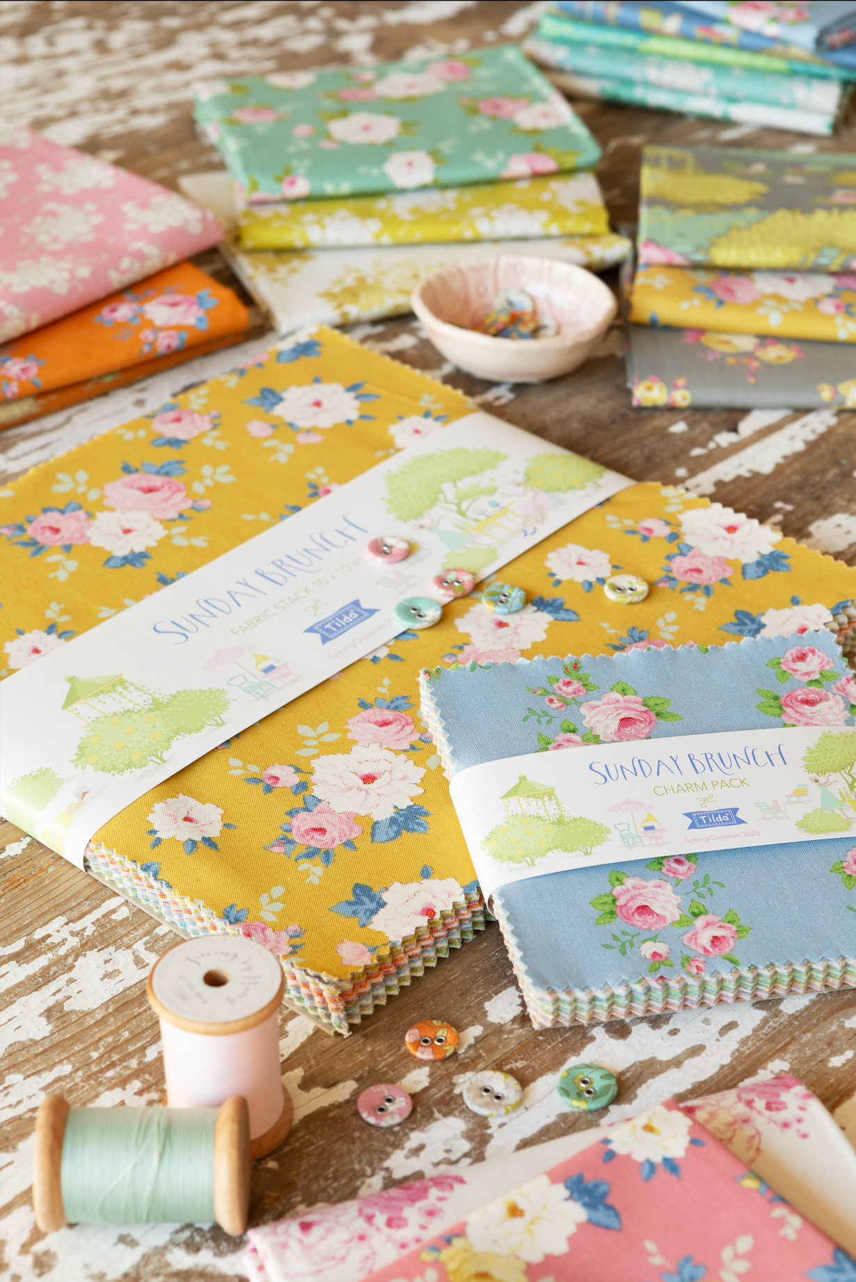 A styled flat-lay of fabric bundles from Sunday in Paris, featuring floral patterns in yellow, blue, pink, and green on a rustic wooden surface with sewing props like spools of thread and buttons.
Photo by Tildafabrics.com [additional_image_link]