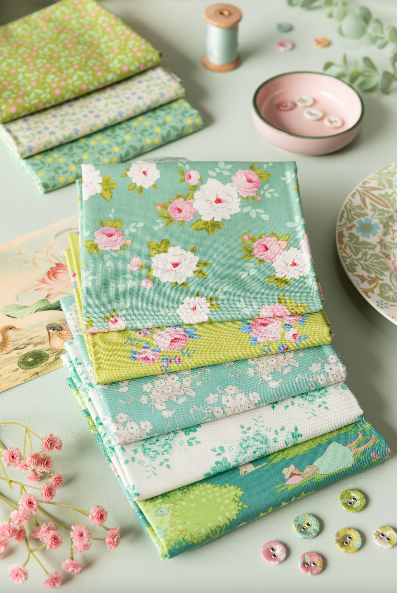 A styled flat-lay of folded fabric pieces with floral and botanical prints in soft pastel tones, arranged on a light surface with sewing-themed props including buttons, a pink dish, thread, and a patterned plate.
Photo by Tildafabrics.com [additional_image_link]