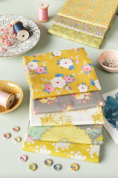A stack of folded fabric pieces with floral and patterned prints in yellow, pink, gray, and white, arranged on a light surface with sewing-themed props like spools, buttons, and a small dish.
Photo by Tildafabrics.com [additional_image_link]