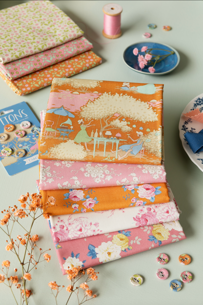 A stack of folded fabric pieces with floral and illustrated prints in pink, orange, cream, and blue, arranged on a light surface with sewing props including buttons, a spool of pink thread, a small dish, and dried flowers.
Photo by Tildafabrics.com [additional_image_link]