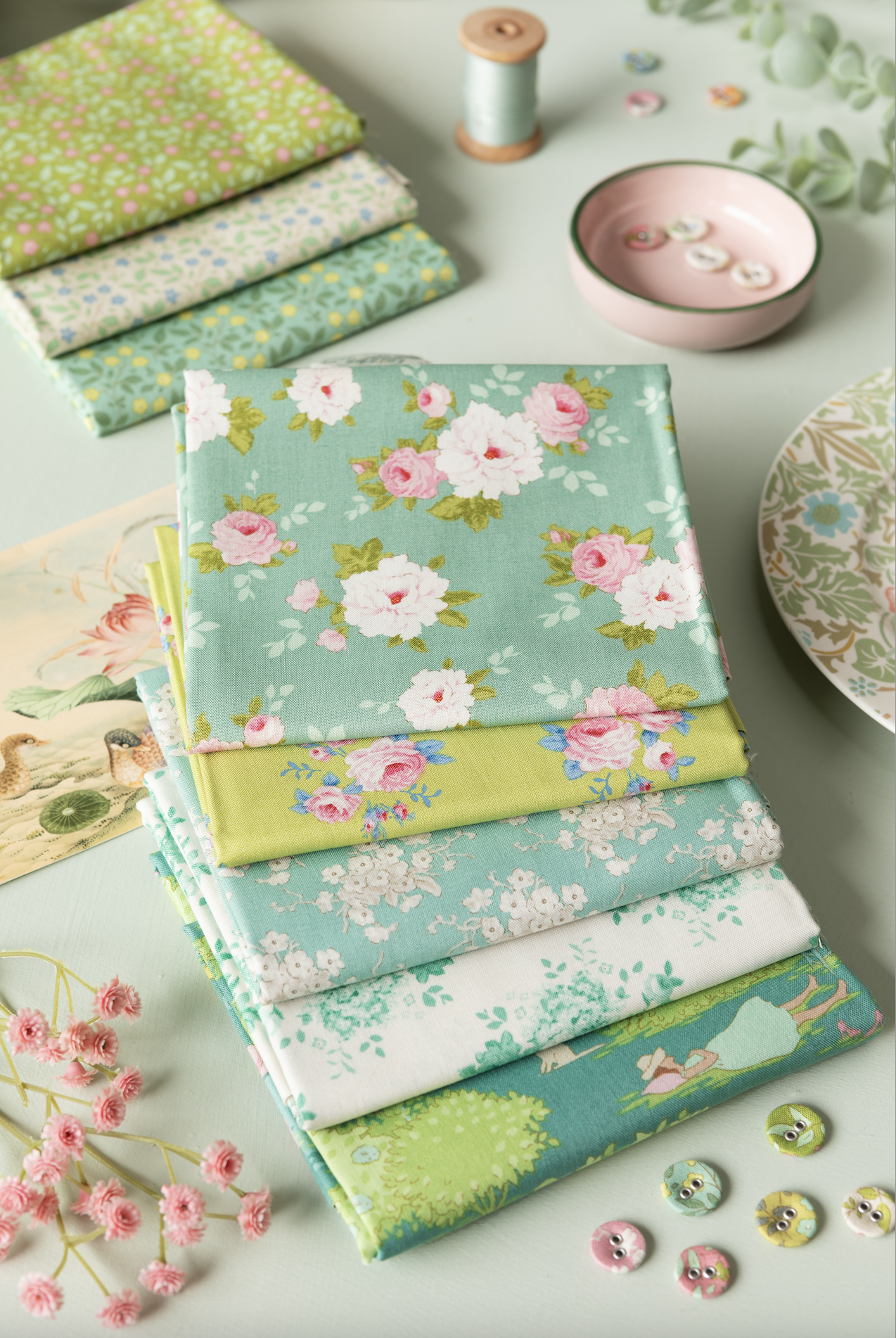 A styled flat-lay of folded fabric pieces with floral and botanical prints in soft pastel tones, arranged on a light surface with sewing-themed props including buttons, a pink dish, thread, and a patterned plate.
Photo by Tildafabrics.com [additional_image_link]