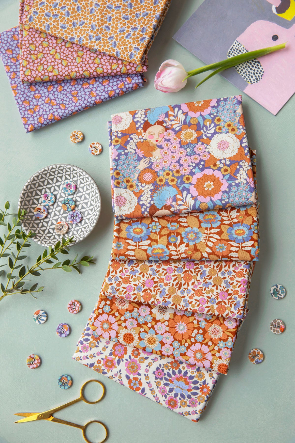 A styled flat-lay of several folded fabric pieces with colorful floral and paisley patterns, arranged on a pale blue-green surface with scissors, buttons, a tulip, and decorative paper props.
Photo by Tildafabrics.com [additional_image_link]