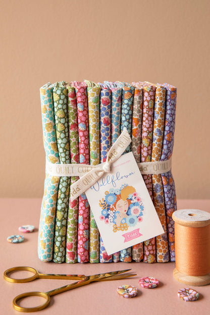 A bundle of colorful patterned fabric rolls tied with a ribbon and card, styled on a pink surface with gold scissors, a spool of peach thread, and small fabric pieces against a warm beige background.
Photo by Tildafabrics.com [additional_image_link]