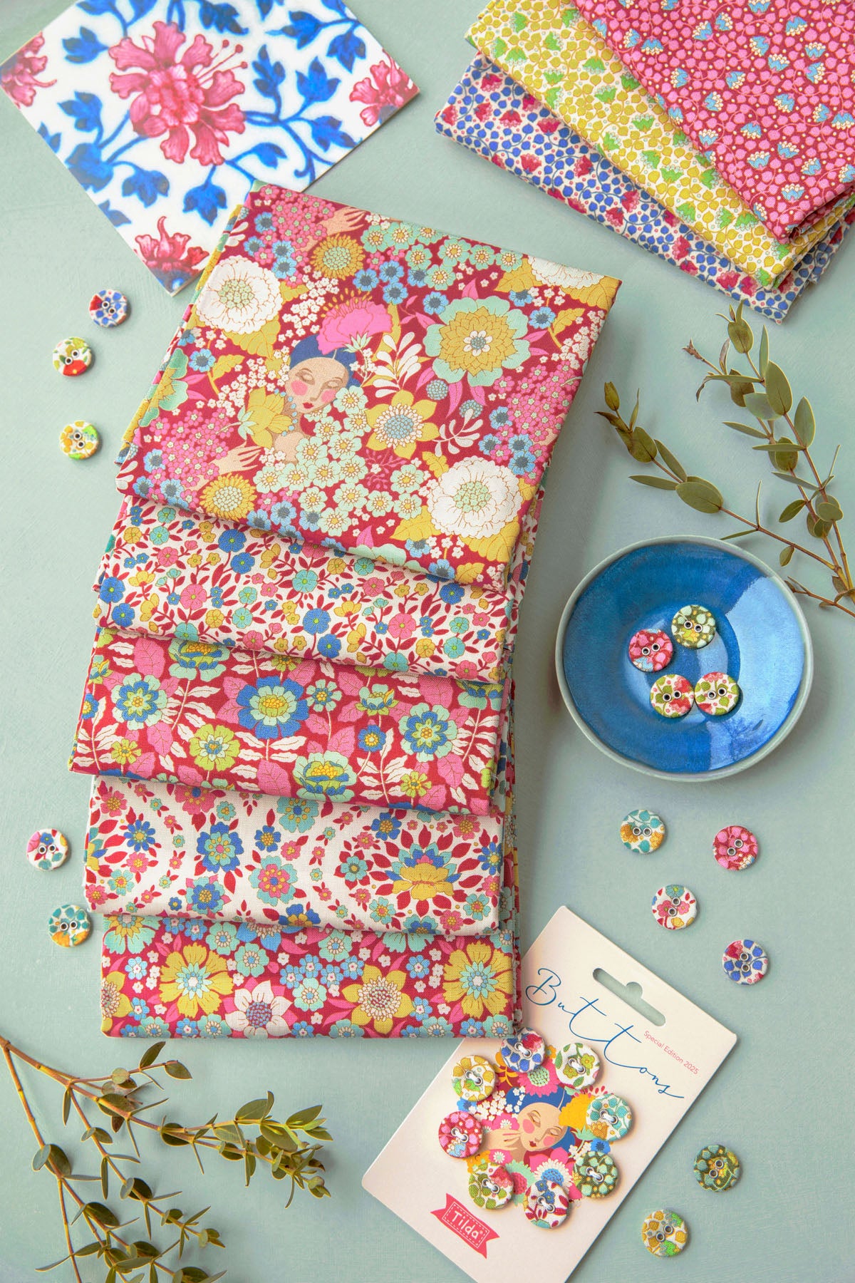 A styled flat-lay of multiple stacks of colorful fabric squares with floral and geometric patterns, alongside loose buttons, a small blue dish, and decorative branches on a pale blue background.
Photo by Tildafabrics.com [additional_image_link]