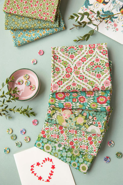 A styled flat-lay of multiple stacks of patterned fabric squares in floral and paisley designs, arranged on a pale blue-green background with decorative buttons, greenery, a pink candle, and a white card with a red heart motif.
Photo by Tildafabrics.com [additional_image_link]