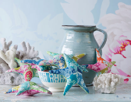 A collection of colorful fabric starfish decorations in various patterns and vibrant colors, arranged around a small white bowl and a rustic teal ceramic pitcher on a light blue surface with coral props.
[additional_image_link]