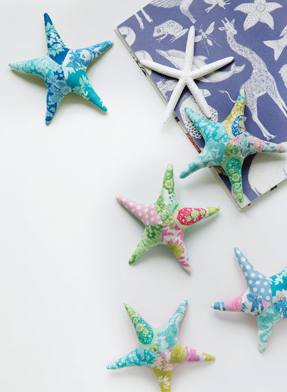 Five fabric starfish ornaments in various colorful floral and polka dot patterns are arranged on a white surface next to a blue patterned book and a white starfish.
[additional_image_link]