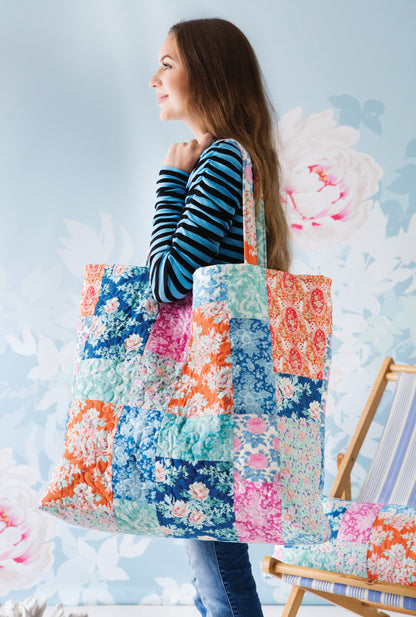 A woman in a blue and black striped shirt carries a large patchwork fabric tote bag with floral prints in blue, pink, orange, and green. She is standing in front of a light blue floral wall, with a wooden chair partially visible on the right.
[additional_image_link]