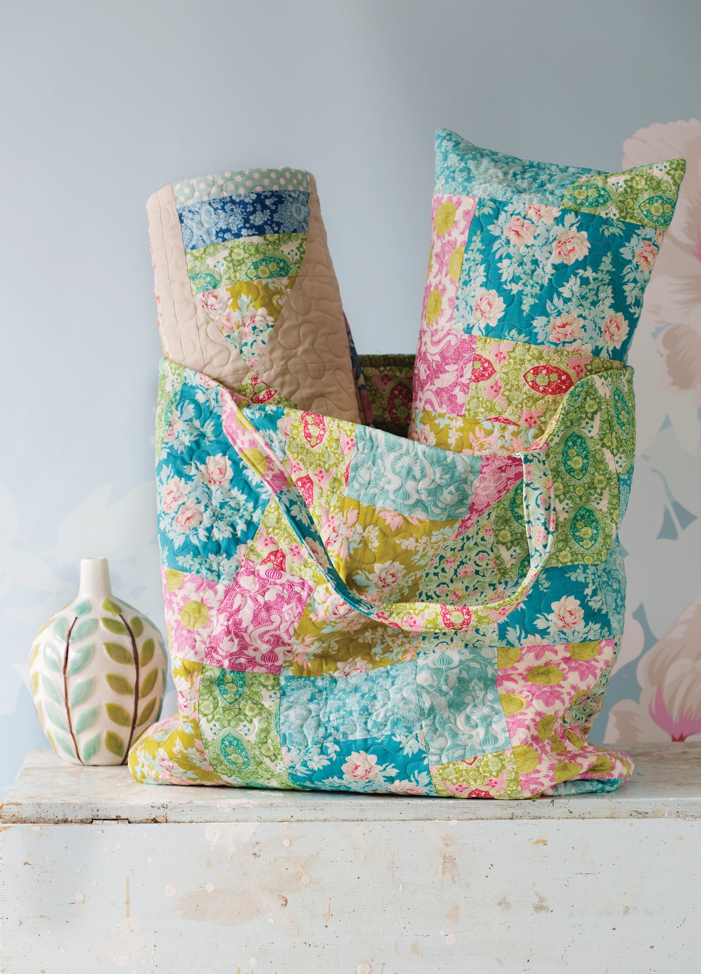 A quilted patchwork tote bag filled with folded quilts, set on a rustic white wooden surface next to a decorative ceramic vase against a soft light blue background.
[additional_image_link]