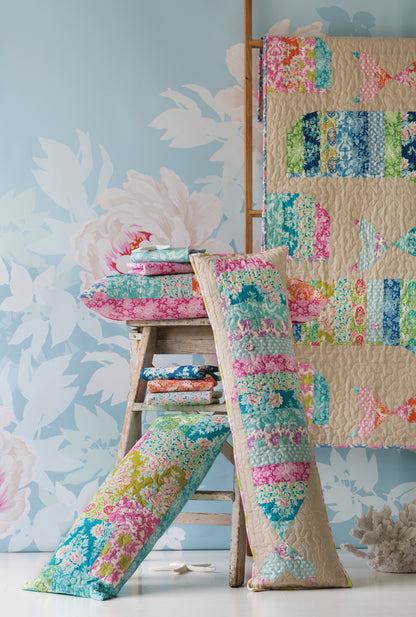 A colorful quilted textile set displayed in a styled room scene, including a patchwork quilt hanging on a wooden stand, two long lumbar pillows, and folded matching fabrics on a rustic stool against a pale aqua floral wall.
[additional_image_link]