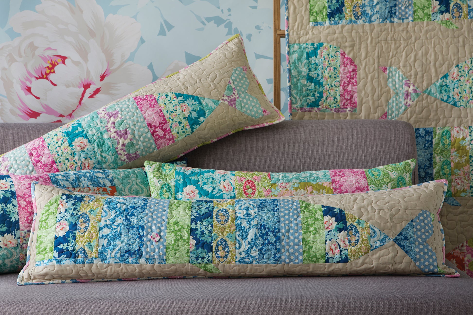 A set of patchwork fish-shaped lumbar pillows in shades of blue, green, pink, and beige, styled on a gray sofa with a matching patchwork quilt in the background.
[additional_image_link]
