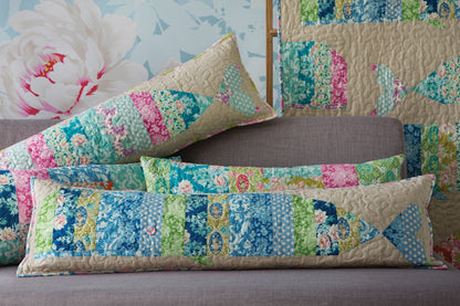 A set of patchwork fish-shaped lumbar pillows in shades of blue, green, pink, and beige, styled on a gray sofa with a matching patchwork quilt in the background.
[additional_image_link]
