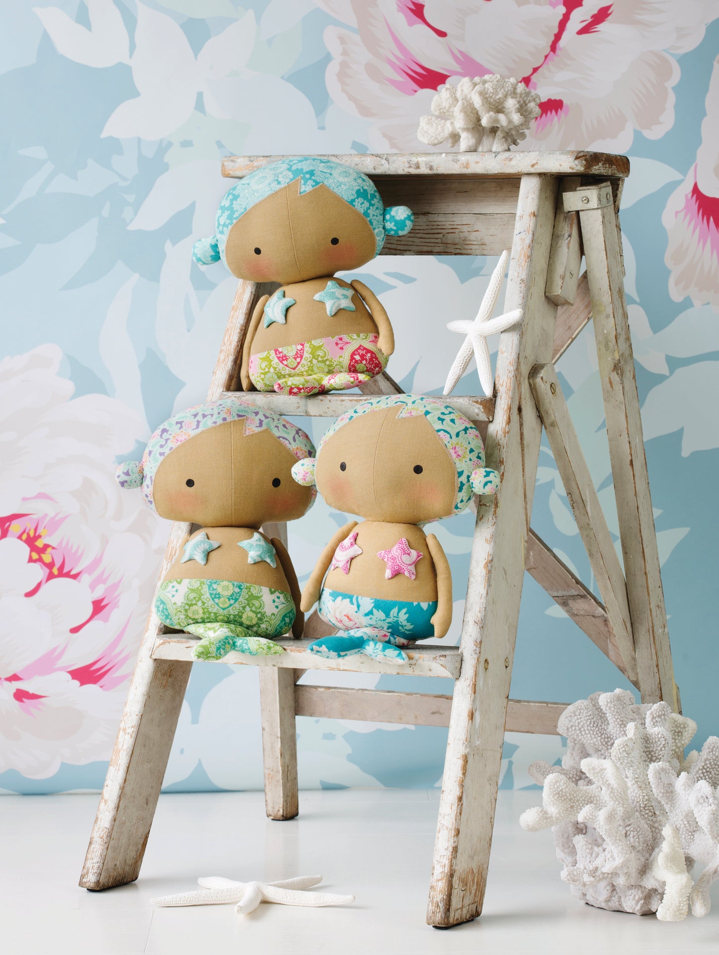 Three small mermaid dolls with tan faces and bodies, mint green hair, and colorful starfish-themed outfits, arranged on a rustic white wooden ladder with coral props against a light aqua floral background.
[additional_image_link]