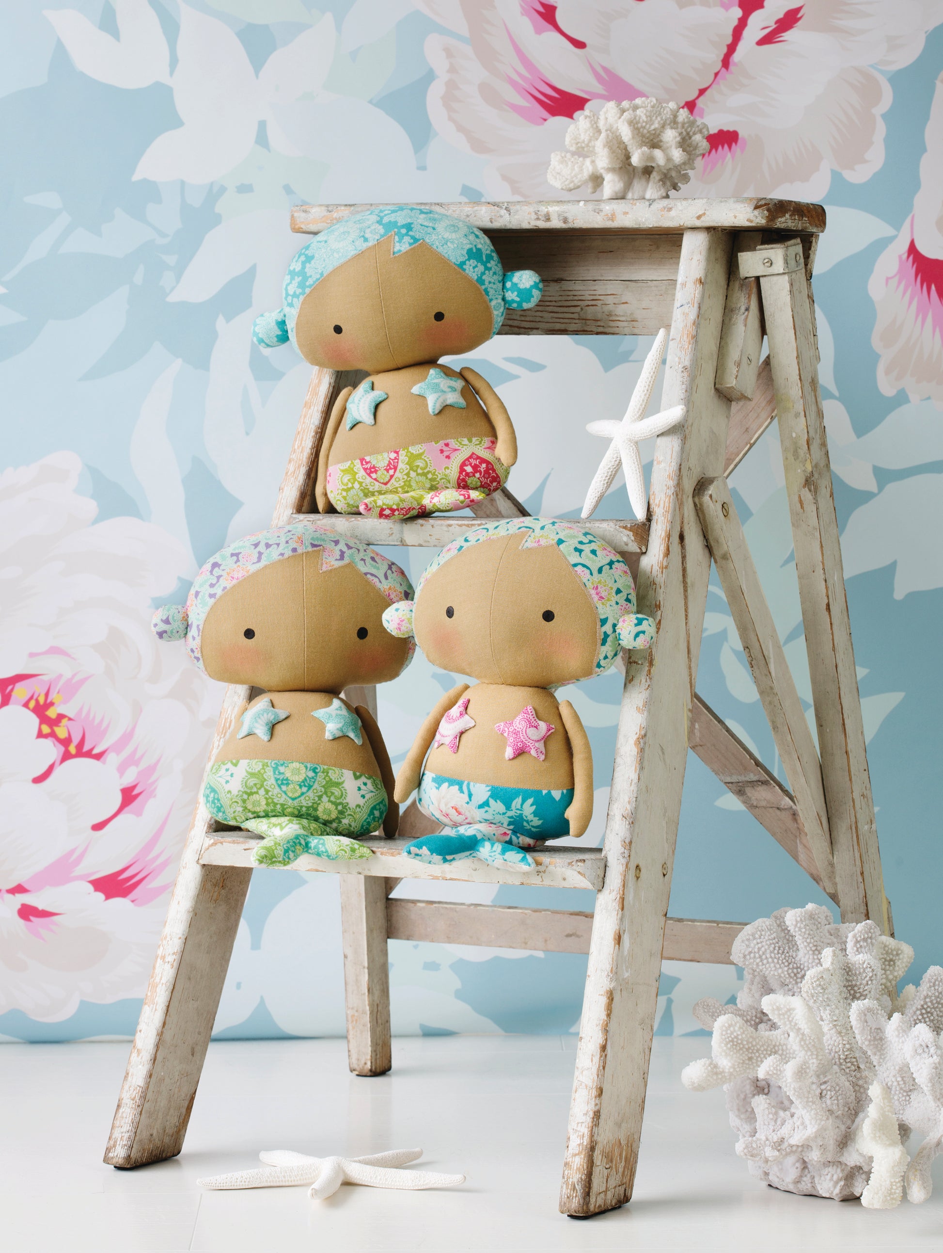 Three small mermaid dolls with tan faces and bodies, mint green hair, and colorful starfish-themed outfits, arranged on a rustic white wooden ladder with coral props against a light aqua floral background.
[additional_image_link]