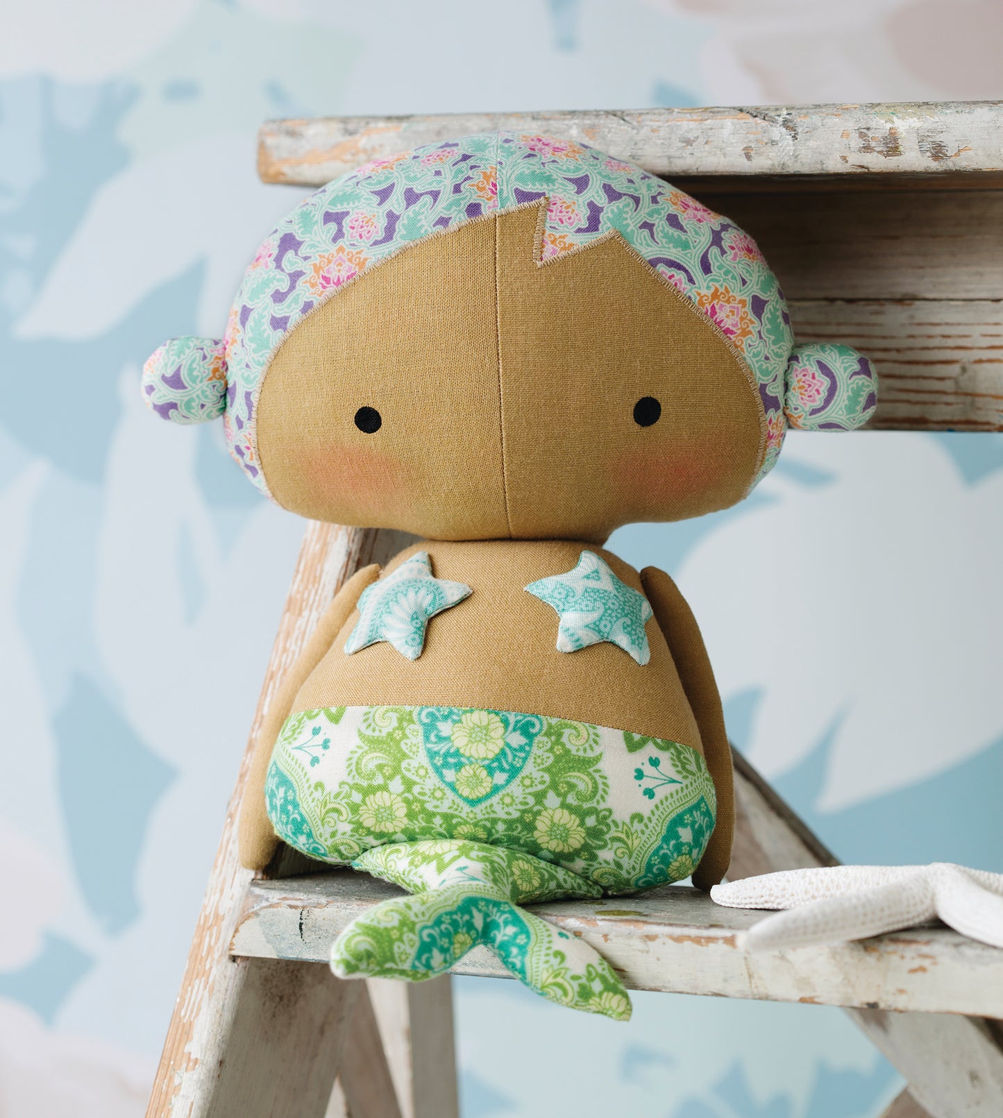 A handmade fabric mermaid doll with a brown body, green patterned hair, and a green patterned tail, sitting on a rustic wooden stool.
[additional_image_link]