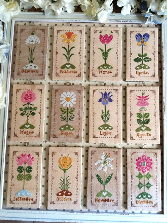 A framed decorative grid showing twelve embroidered floral cards, each labeled with a different month in Italian, arranged in a 3x4 layout inside a white wooden frame with white flowers around the border.
Photo by CuoreBatticuore.com 
[image_link]
