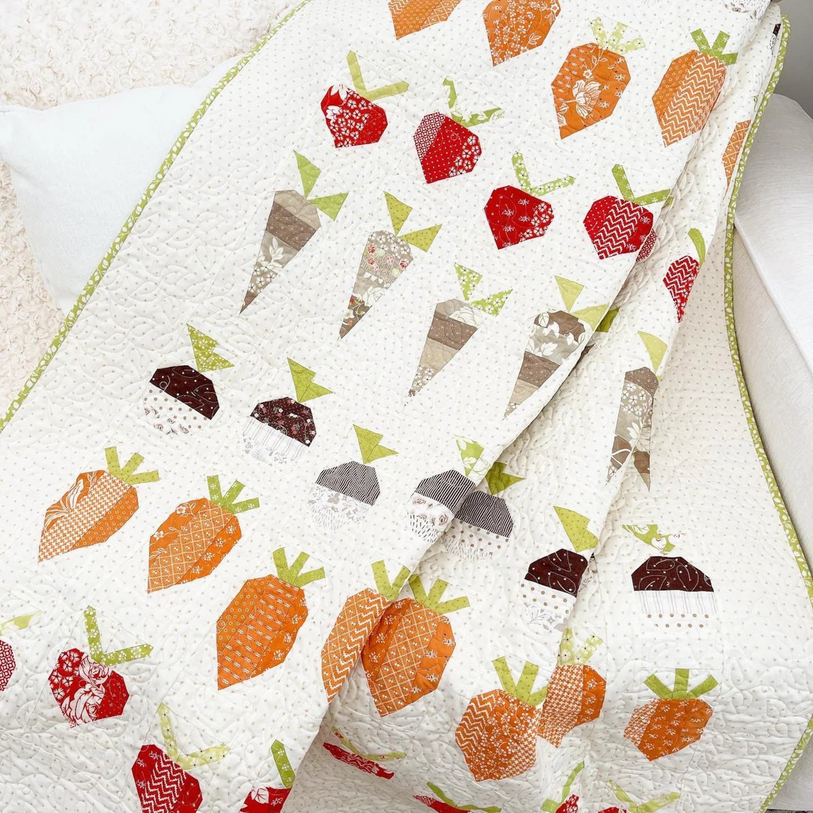 A close-up of a quilted textile featuring a pattern of stylized carrots and apples in orange, red, brown, gray, and green on an off-white background, with a light green stitched border.
Photo by ThePatternBasket.com [additional_image_link]