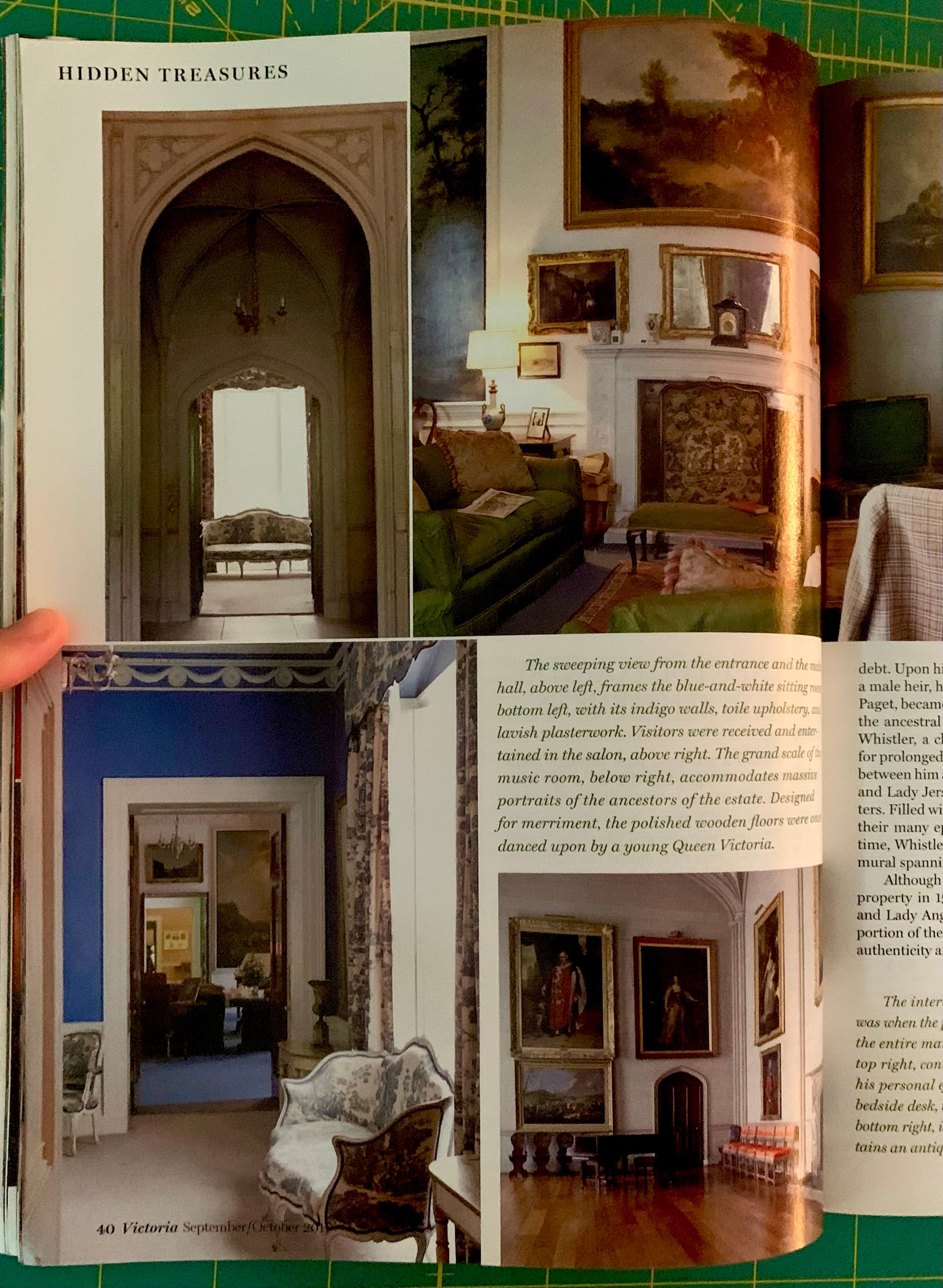 An open book or magazine spread showing interior architectural photographs and text, photographed from below on a green cutting mat with a hand holding the left page edge.
[additional_image_link]