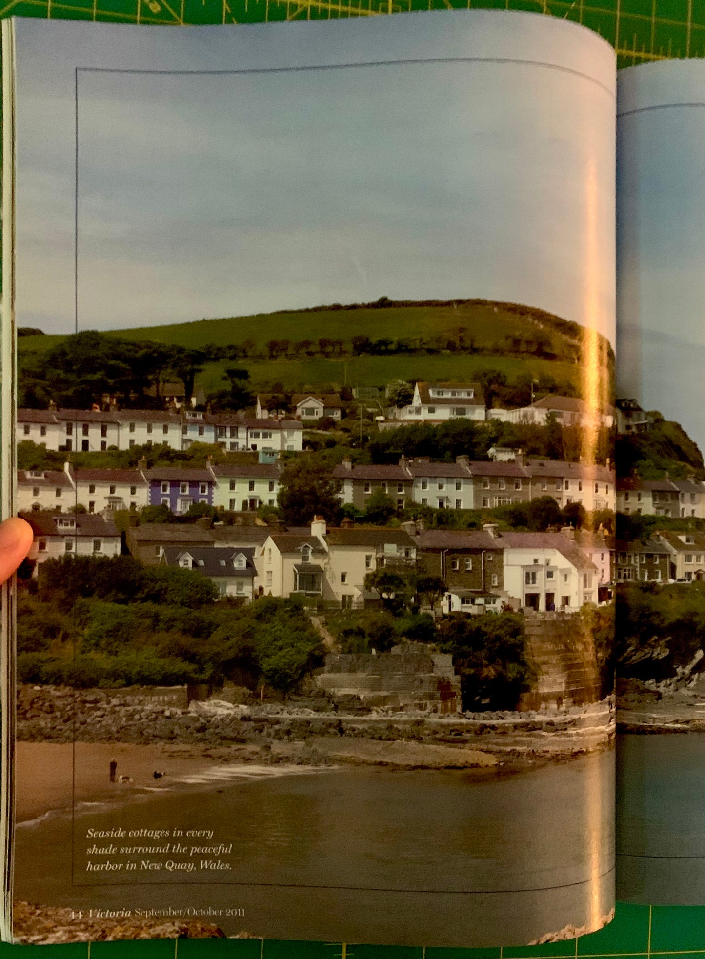 An open book or magazine page showing a coastal village scene with houses built into a green hill above a rocky shoreline and water. A hand is holding the left edge of the page, and the page is photographed against a green cutting mat background.
[additional_image_link]