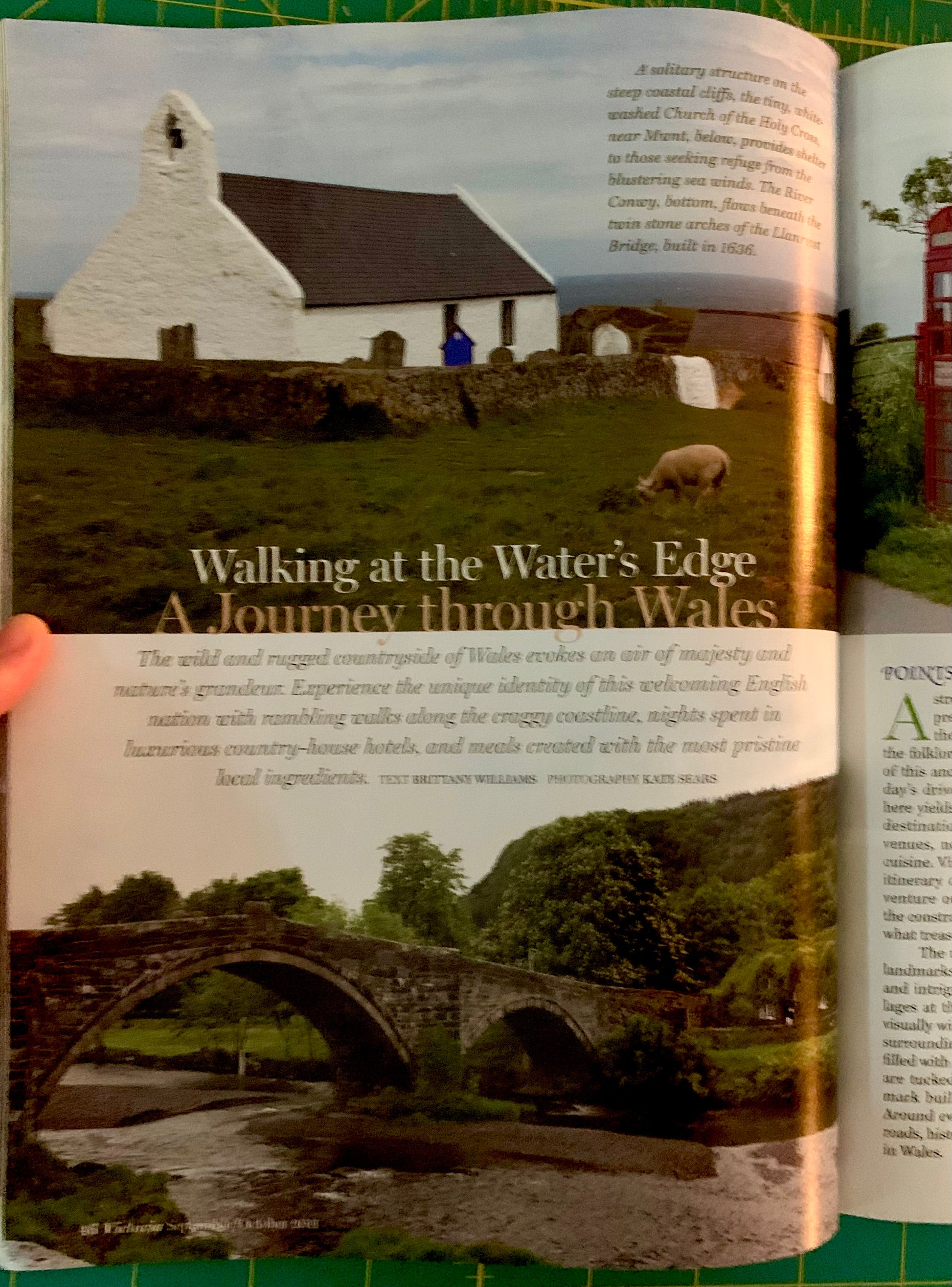 An open printed magazine or book spread showing a travel article titled 'Walking at the Water's Edge: A Journey through Wales,' with landscape photographs of a white church, a sheep on a field, and stone bridges over water. A human hand is visible holding the left page edge.
[additional_image_link]