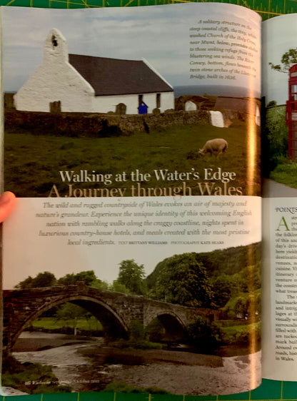 An open printed magazine or book spread showing a travel article titled 'Walking at the Water's Edge: A Journey through Wales,' with landscape photographs of a white church, a sheep on a field, and stone bridges over water. A human hand is visible holding the left page edge.
[additional_image_link]