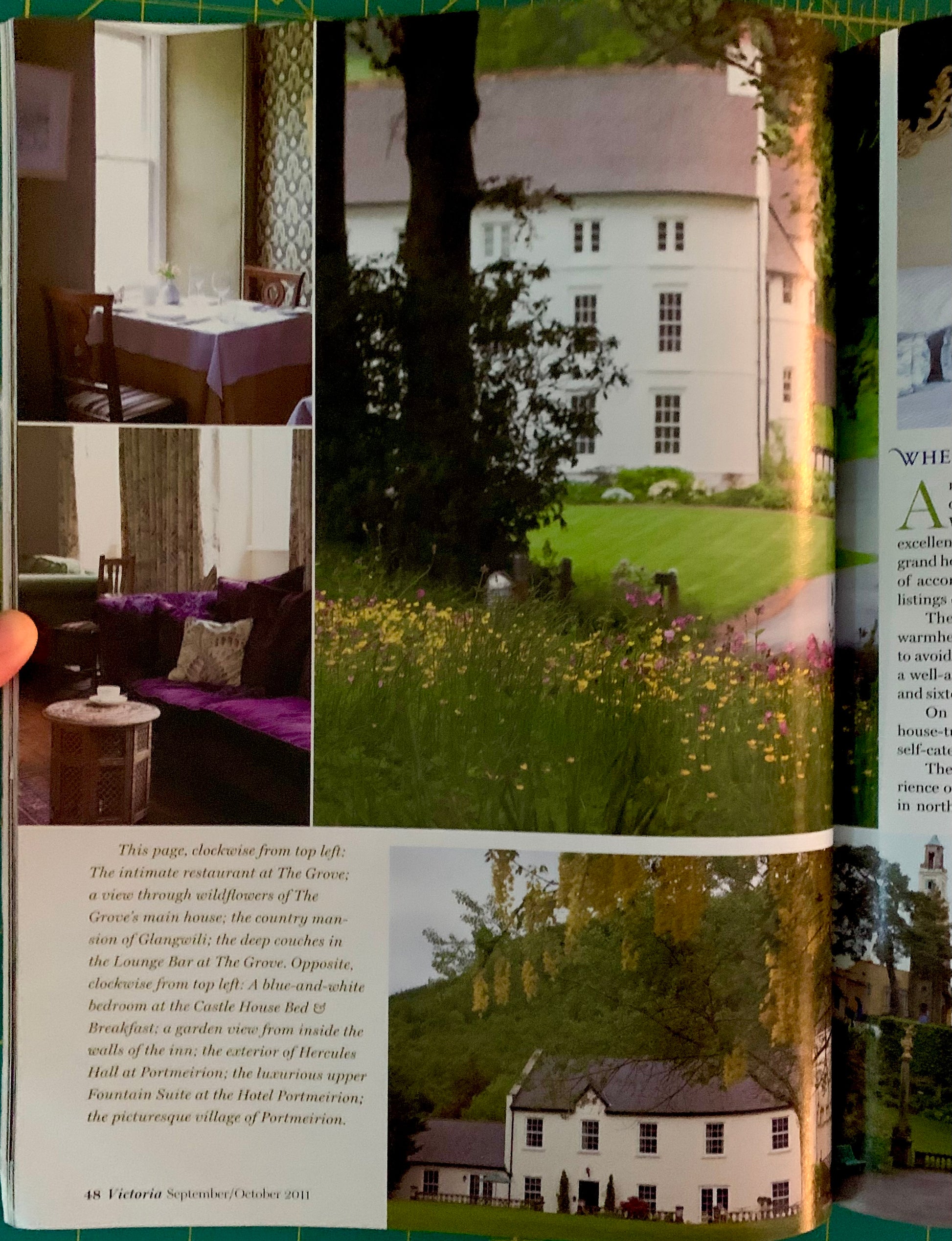 An open magazine or book page showing a large outdoor photograph of a white house with a green lawn and trees, plus smaller inset photos and text blocks. A human finger is visible holding the page open on the left edge.
[additional_image_link]