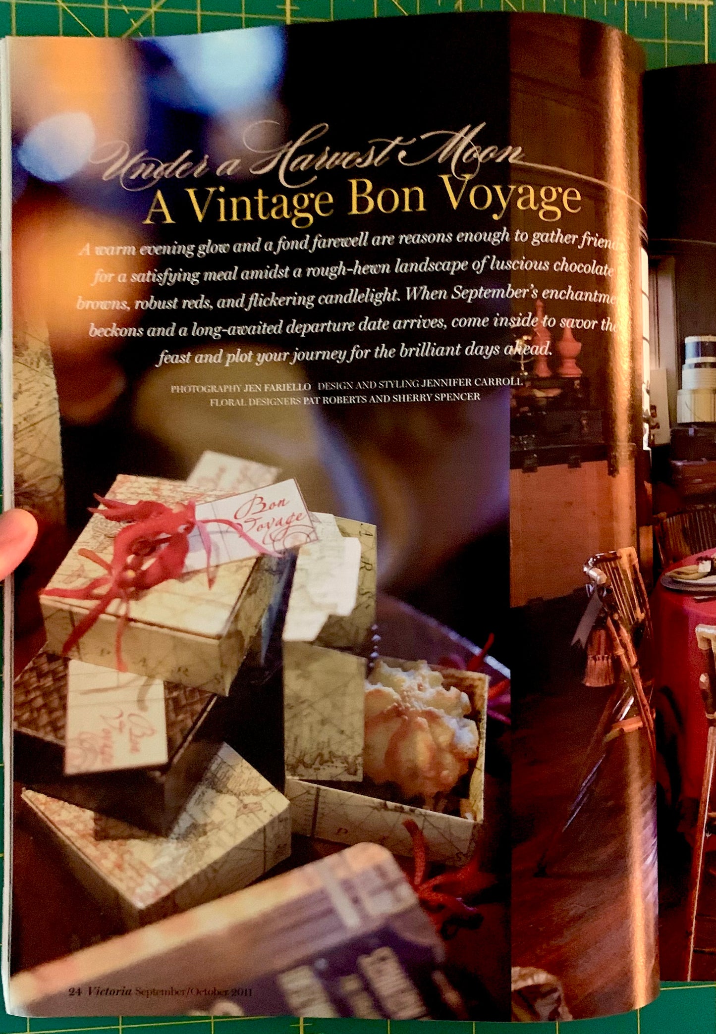 An open magazine or book spread featuring a travel-style article titled 'Hares a Harvest Moon A Vintage Bon Voyage' with a photo of wrapped gift boxes tied with red ribbons on a table. A hand is holding the left page edge, and the right page shows a warm interior scene with candles and decor.
[additional_image_link]