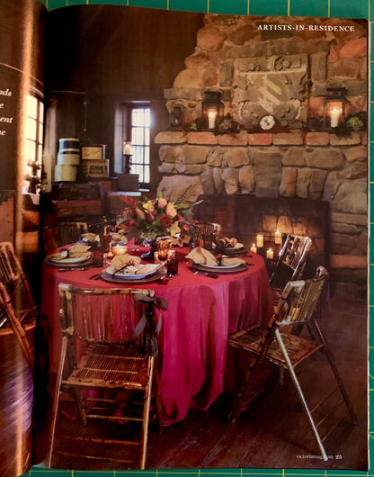 A printed interior scene showing a rustic dining room with a round table covered in a bright pink tablecloth, set with plates, candles, and chairs in front of a large stone fireplace. The photo appears inside an open book or magazine, with green grid lines visible around the edges.
[additional_image_link]