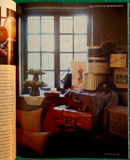 An open book or magazine spread showing a vintage interior scene with a large window, stacked suitcases, hats, framed art, and furniture. The right page has the text 'ARTISTS-IN-RESIDENCE' at the top.
[additional_image_link]