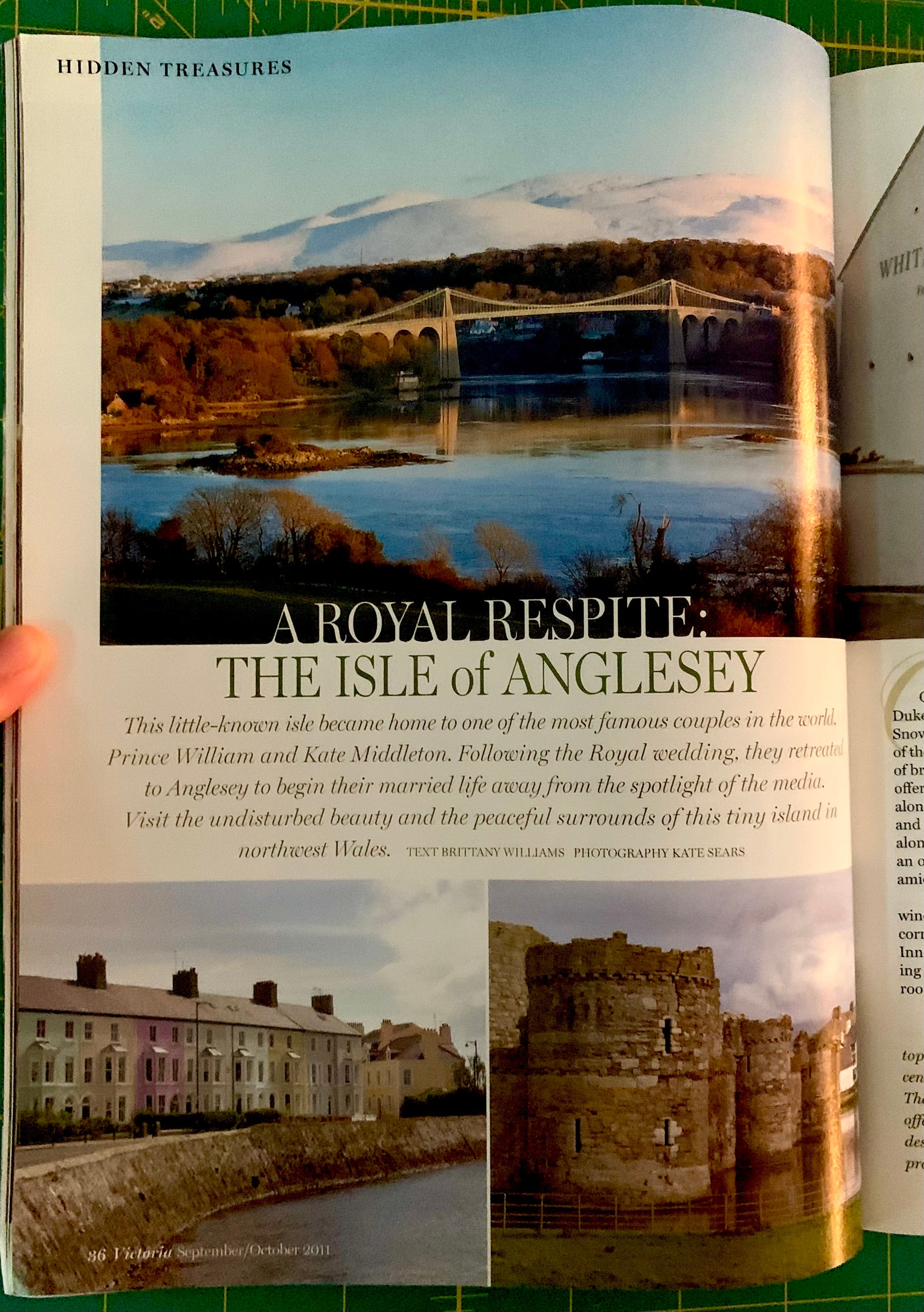 An open book or magazine page featuring a landscape photograph of Anglesey with a bridge over water, a caption, and smaller architectural and ruin photos. A human finger is visible holding the page open.
[additional_image_link]
