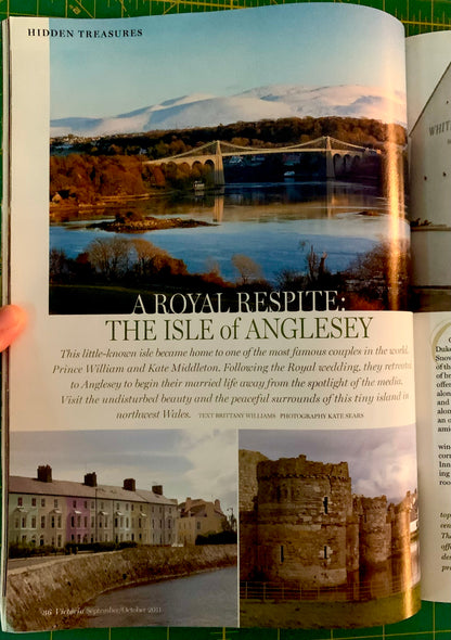 An open book or magazine page featuring a landscape photograph of Anglesey with a bridge over water, a caption, and smaller architectural and ruin photos. A human finger is visible holding the page open.
[additional_image_link]