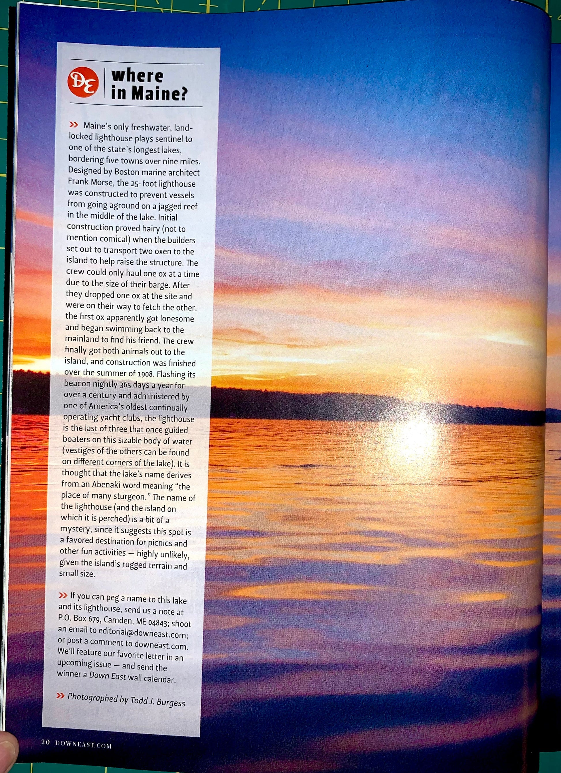An open magazine or book page showing a scenic photograph of a lighthouse over water on the right and a text column headed 'Where in Maine?' on the left. A hand is holding the page at the bottom left.
[additional_image_link]