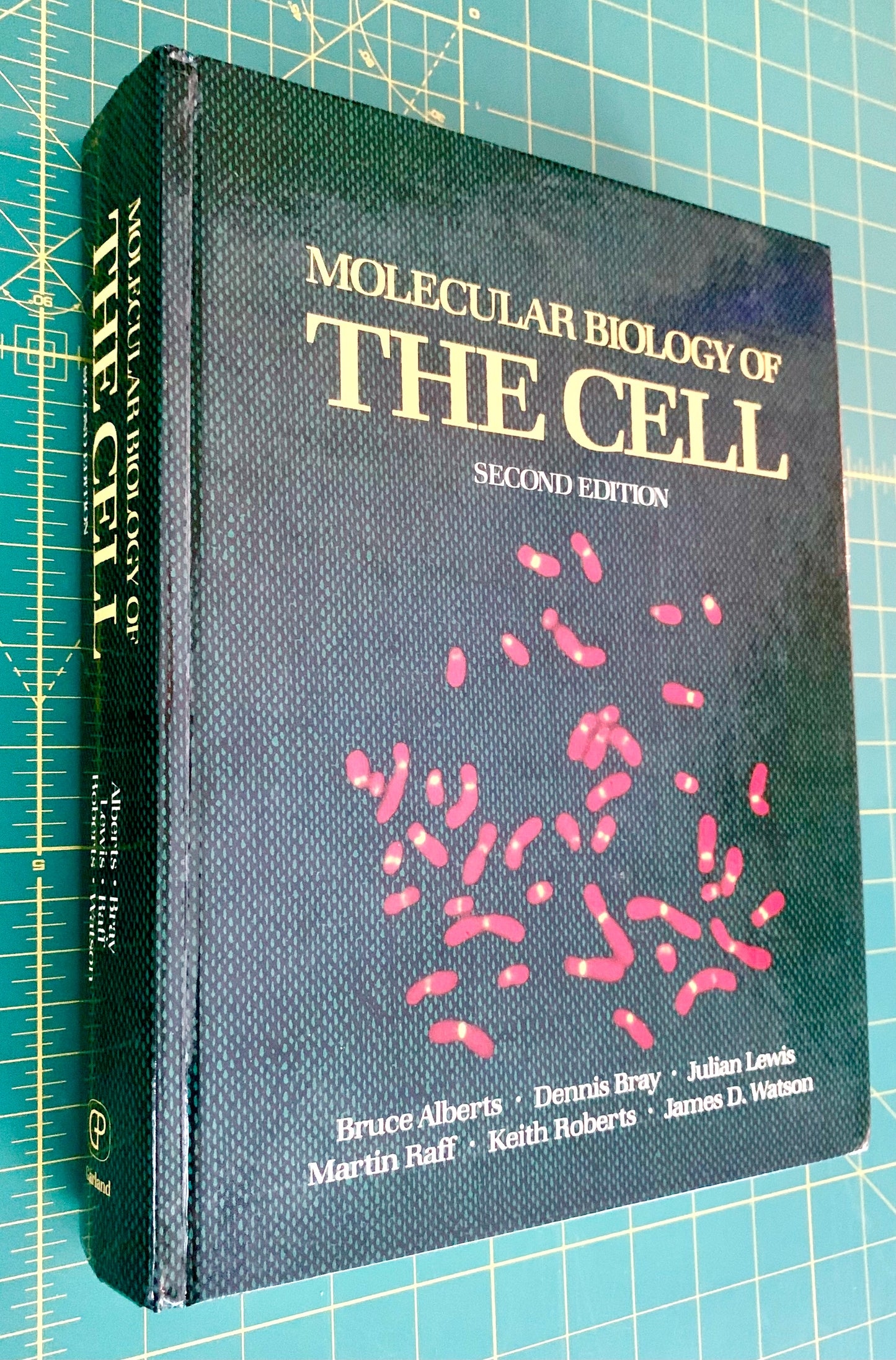 A hardcover textbook titled 'Molecular Biology of the Cell, Second Edition' lying on a green cutting mat with grid lines. The cover is dark textured black with gold title text and pink cellular illustrations.
[additional_image_link]