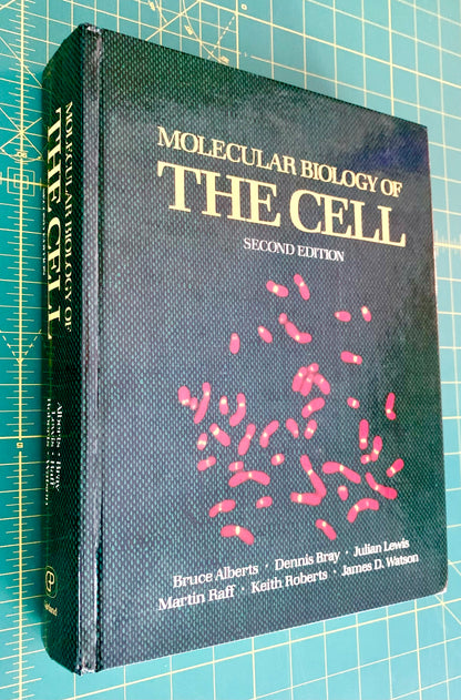 A hardcover textbook titled 'Molecular Biology of the Cell, Second Edition' lying on a green cutting mat with grid lines. The cover is dark textured black with gold title text and pink cellular illustrations.
[additional_image_link]