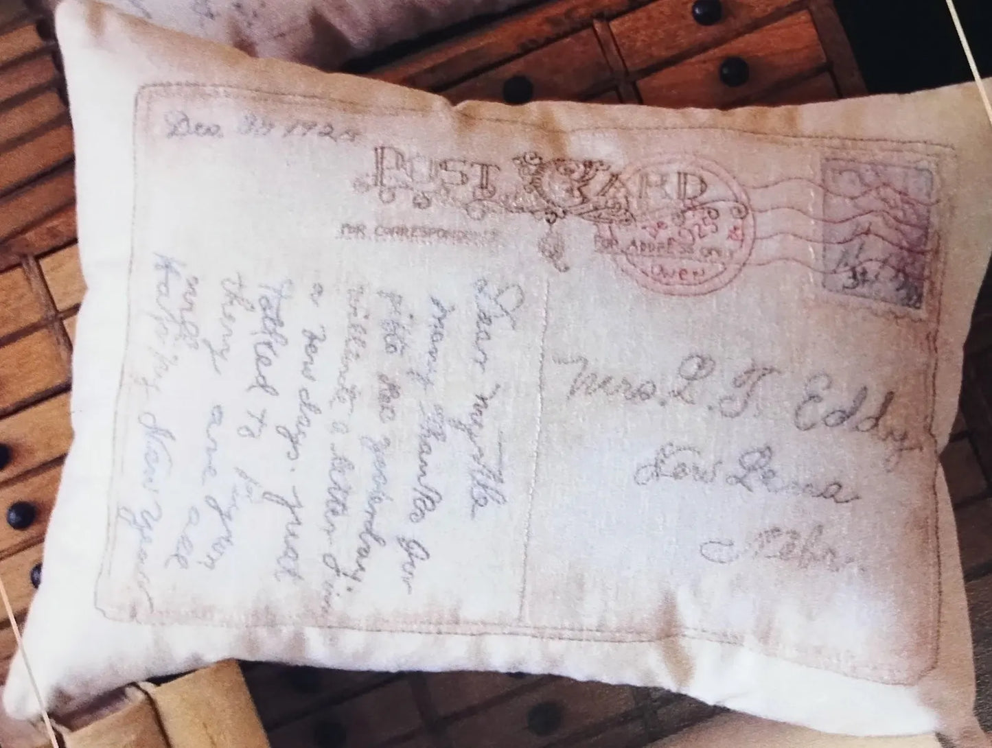 A square decorative throw pillow featuring an antique postcard design with cursive handwriting and a red postage stamp motif, resting on a rustic wooden surface.
Photo by SamSarah Studio Designs [image_link]