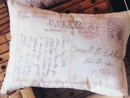 A square decorative throw pillow featuring an antique postcard design with cursive handwriting and a red postage stamp motif, resting on a rustic wooden surface.
Photo by SamSarah Studio Designs [image_link]