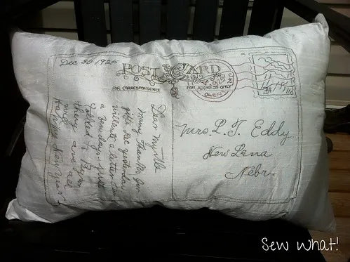 A white fabric lumbar pillow with a vintage postcard design, featuring cursive handwriting and a stamp motif, resting on a dark wooden chair.
Photo by SamSarah Studio Designs [additional_image_link]