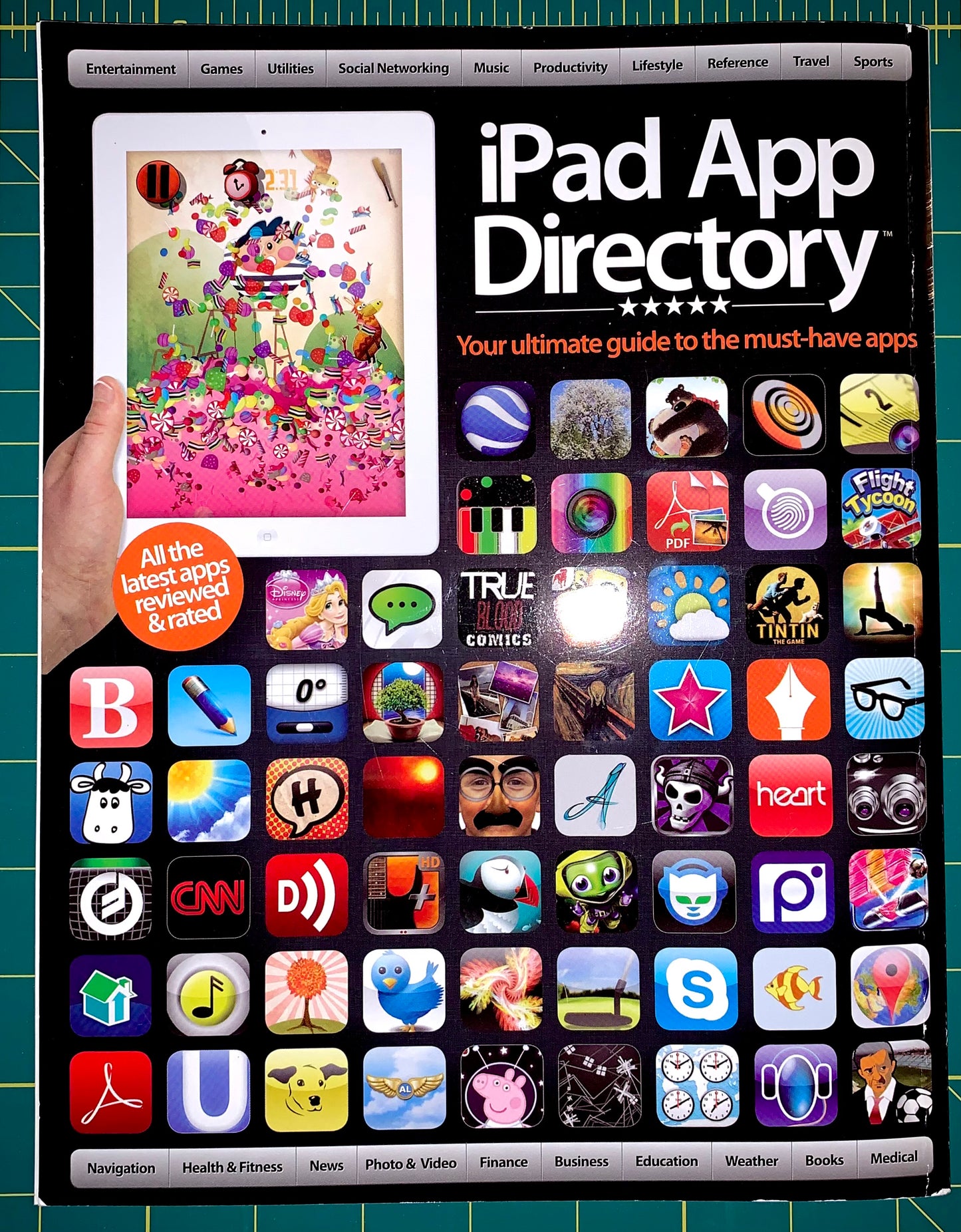 An iPad App Directory book or guide shown flat on a green cutting mat, featuring a large iPad screen with a colorful illustration and a grid of many app icons. A hand is visible holding the left edge of the iPad.
[additional_image_link]